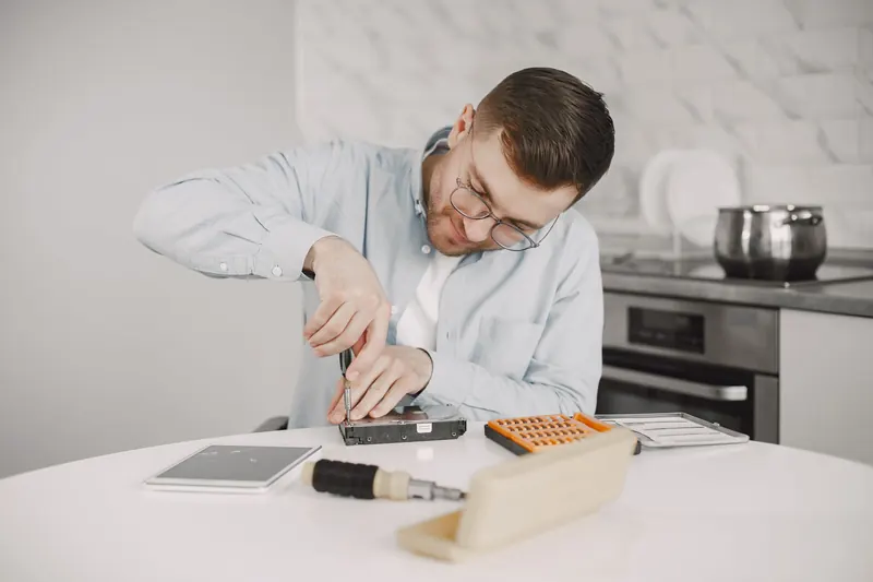 Técnico con lentes desarma con un destornillador una pieza electrónica sobre una mesa, con sus herramientas al lado